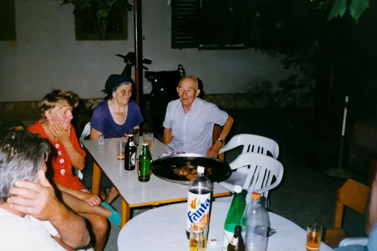 Slika prikazuje babu i didu kako sjede s turistima nakon obroka – trenutak opuštanja, smijeha i druženja. U kadru se vidi svojevrsni mix vremena: prve plastične stolice s terasa, tada masovno kupovane u Italiji, stoje uz stare drvene stolice i čvrsti drveni stol, koji se i danas koristi u kuhinjici konobe apartmana An- Mar. Nije se pazilo na estetiku, već na ono što je bilo važno – da se ljudi okupe, podruže, zapjevaju i podijele obrok. Svaki predmet nosi trag vremena i priču o dugovječnosti, jednostavnosti i pravim vrijednostima nekadašnjeg turizma.