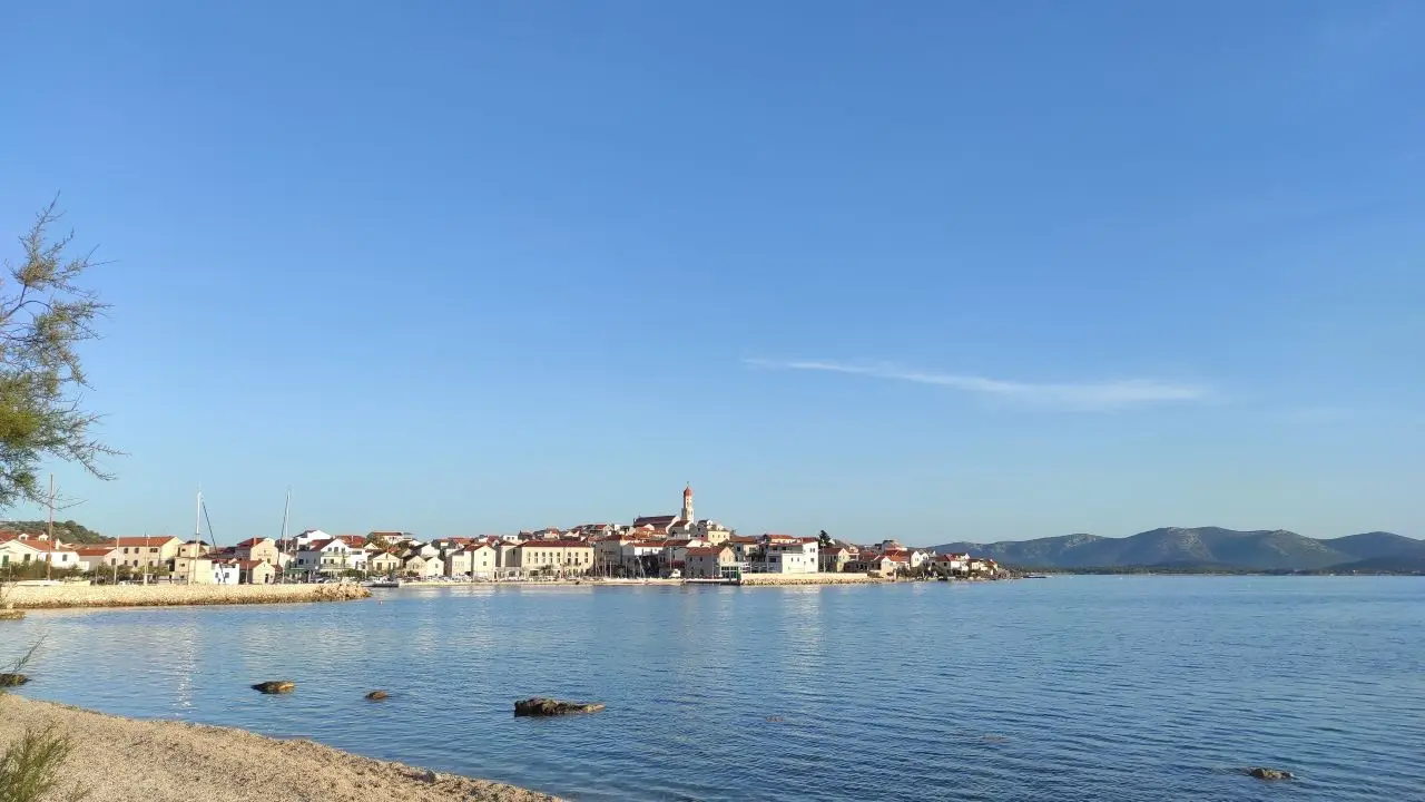 Što posjetiti 1 Slika prikazuje pogled na mjesto Betina na otoku Murteru, snimljen s plaže u blizini parkirališta. U kadru se riva s privezanima tradicionalnim drvenim brodicama – gajetama – koje su dio lokalne pomorske baštine. U pozadini se uz more proteže staro naselje s karakterističnom dalmatinskom arhitekturom. Ova scena dočarava spoj tradicije, mirnog priobalnog života i autentične mediteranske atmosfere.
